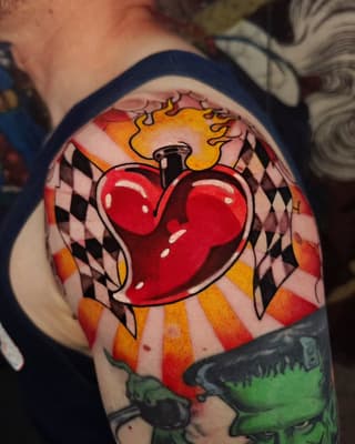 Sacred heart tattoo with flames, harlequin pattern background, and green element below on upper arm in red, yellow, orange, and black.