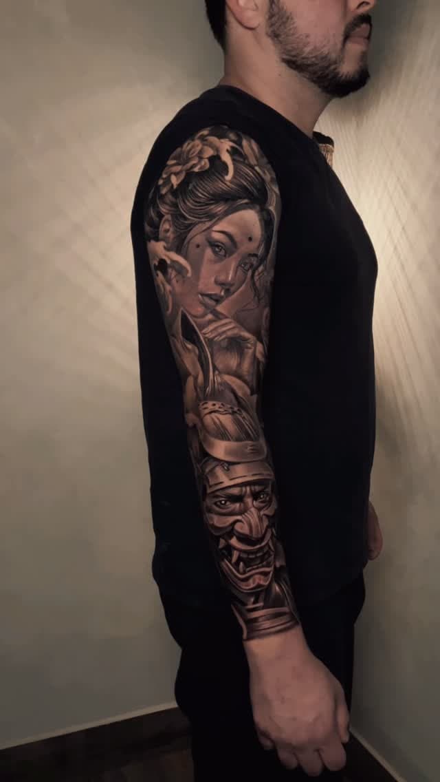 Man's arm displays a full-sleeve tattoo featuring a woman's portrait, demon mask, and decorative elements in black and gray ink.