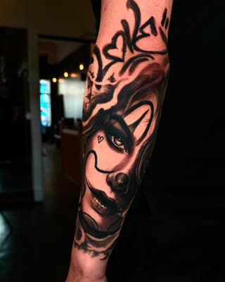 Demon face tattoo on the forearm with detailed shading, sharp facial features, horns, and a small heart marking near the eye in black ink.
