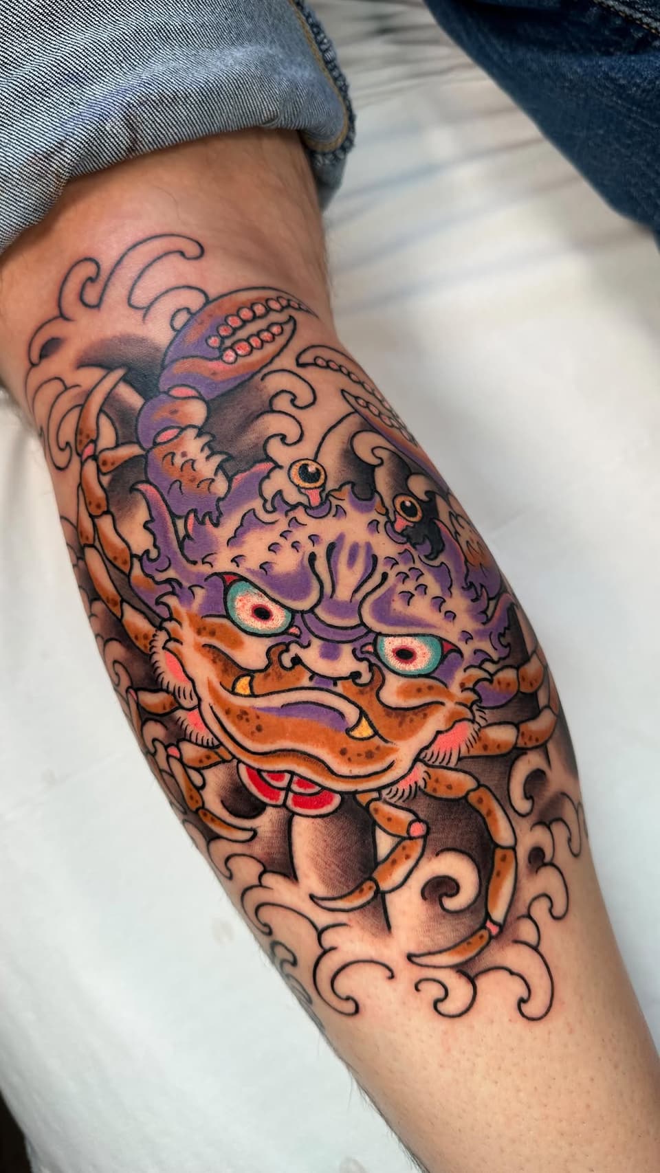 Colorful oni mask tattoo on the forearm featuring orange and purple tones with ornate swirling details, red mouth, and piercing eyes.