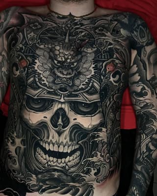 Large chest and sleeve tattoo featuring an ornate demon mask above a skull with decorative scrollwork and floral elements in black and grey ink.