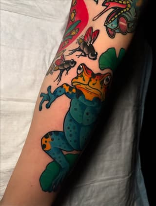 Colorful arm tattoo featuring a spotted lizard with orange and blue markings, surrounded by insects and foliage in green and red tones.