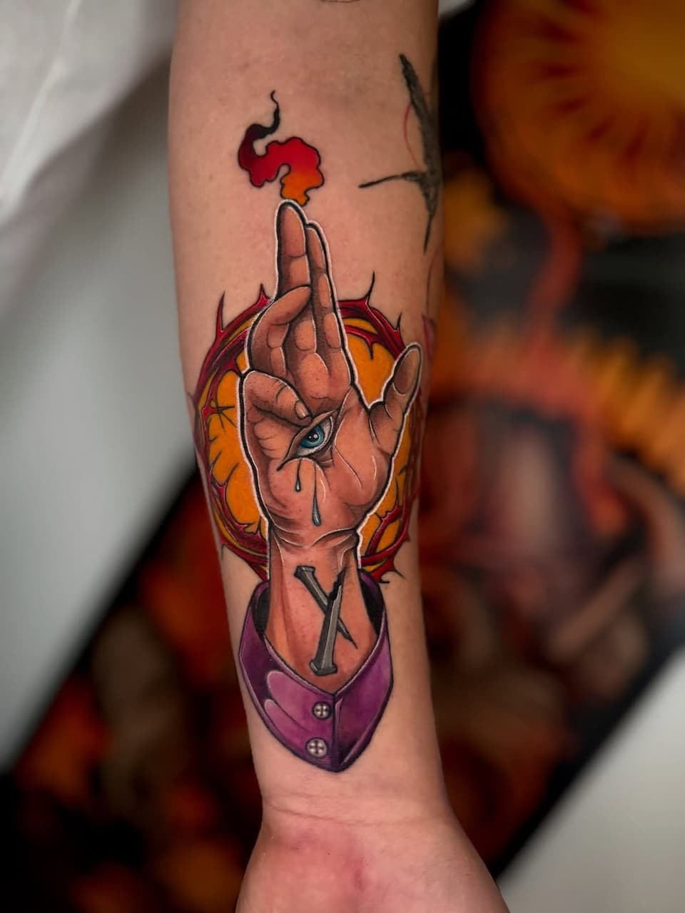Colorful forearm tattoo of an anatomical heart with a hand gesture, flames, and a purple pouch below, rendered in red, orange, and yellow tones.