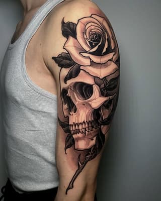 Rose flower and skull design tattooed on upper arm in black ink with peach and tan shading, on a person wearing a gray tank top.