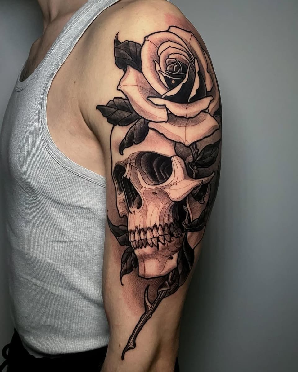 Rose flower and skull design tattooed on upper arm in black ink with peach and tan shading, on a person wearing a gray tank top.