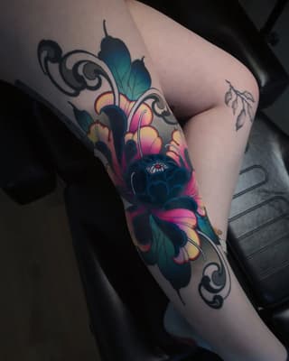 Colorful flower tattoo on the thigh featuring vibrant pink, yellow, and teal petals with ornamental black scrollwork and green leaves.
