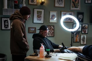 Tattoo artist with visible arm tattoos works on a client's leg under a ring light in a studio with framed artwork on sage green walls.