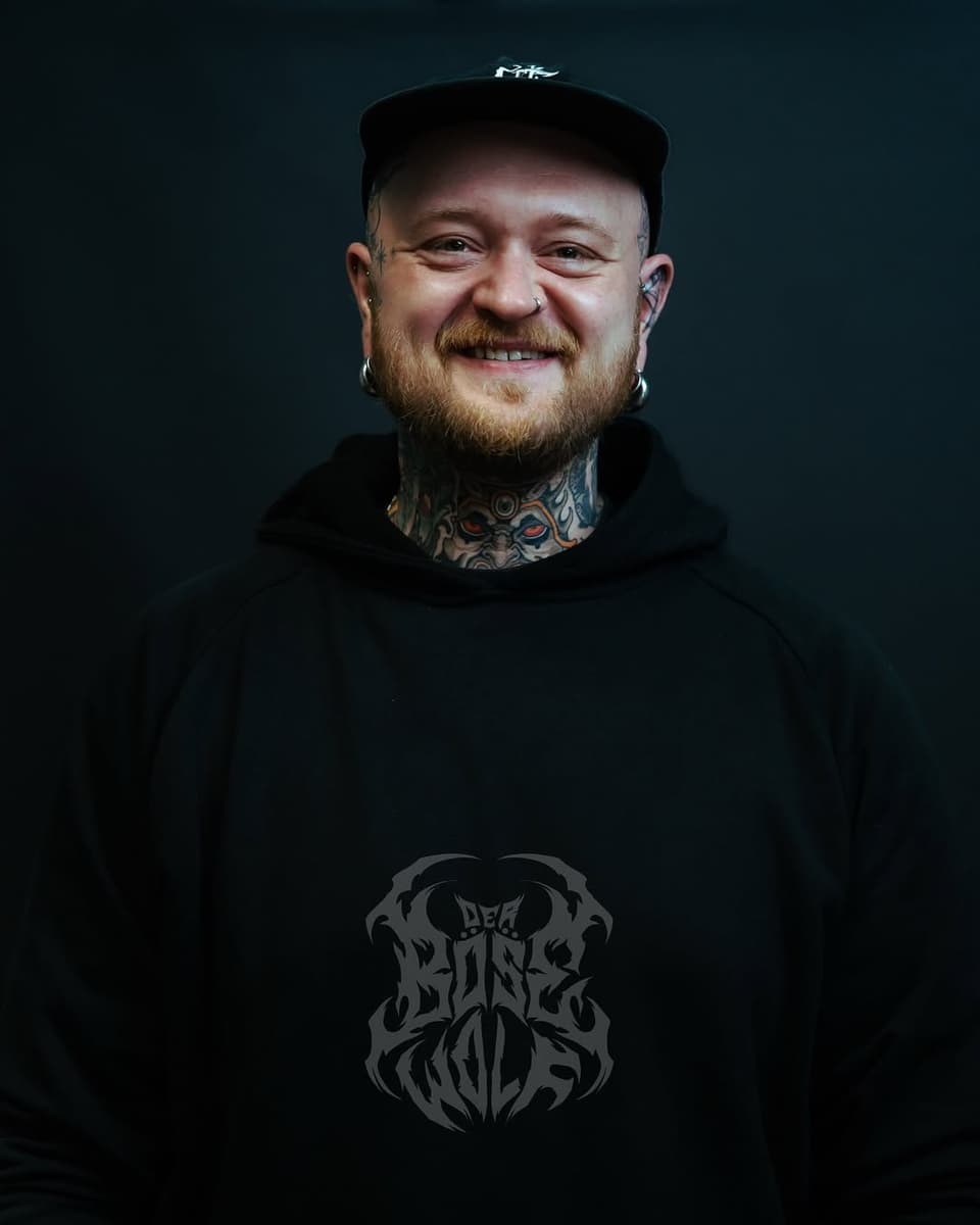 Portrait of a bearded man wearing a black cap and hoodie with an intricate neck tattoo and dark emblem on the chest against a dark background.