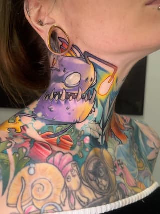 Colorful neck and chest tattoo featuring a purple monster with large eyes, surrounded by vibrant flowers and abstract designs in yellow, pink, green, and blue.