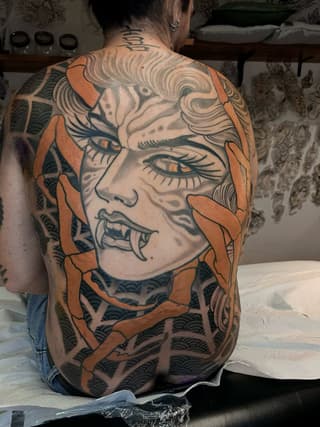 Back tattoo featuring a face with expressive eyes and open mouth, surrounded by orange petal shapes and dark patterned elements.