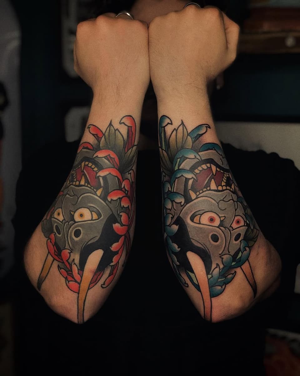 Matching hand tattoos featuring ornate masked faces with leaf motifs, colored in red, teal, and gold on both wrists.