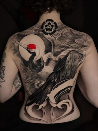 Back tattoo featuring a crane holding a red sun circle, surrounded by wave patterns and ornamental designs in black and gray ink.