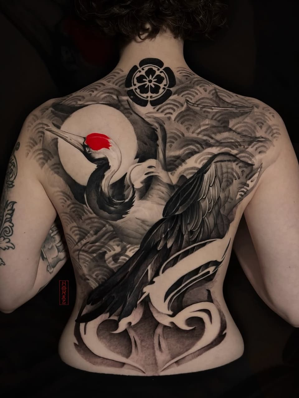 Back tattoo featuring a crane holding a red sun circle, surrounded by wave patterns and ornamental designs in black and gray ink.
