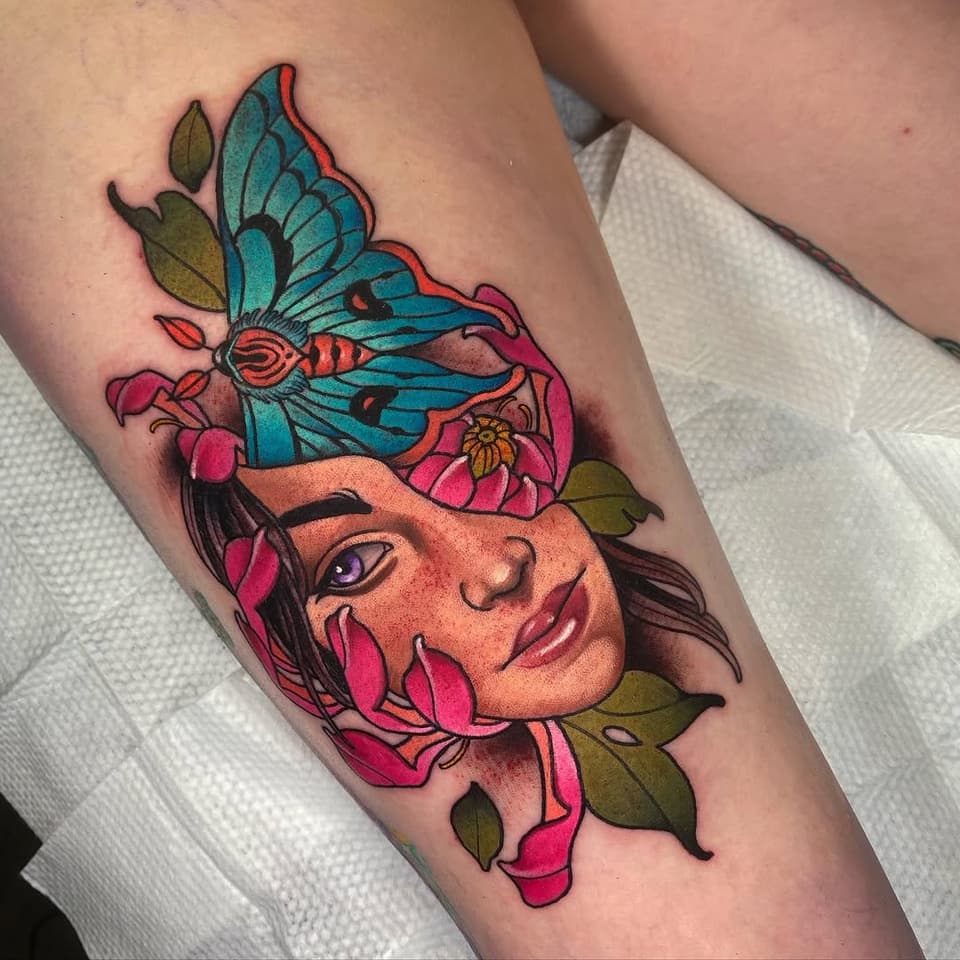 Colorful arm tattoo featuring a woman's face with purple eyes surrounded by magenta flowers, turquoise butterfly, and olive-green leaves.