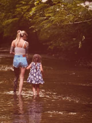 Woman with visible arm tattoo wading in a river with a young child, showing detailed ink work on her shoulder and upper arm in natural daylight.