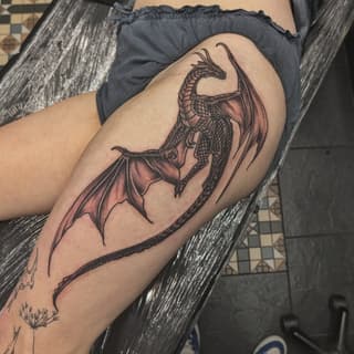 Dragon tattoo on thigh with spread wings and detailed scales in black and red ink, displayed on seated person's leg.