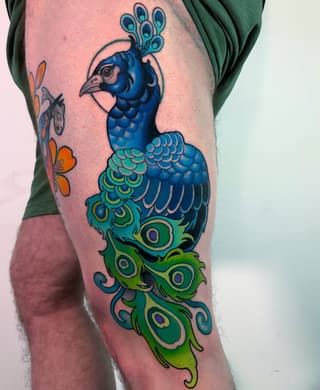 Colorful peacock tattoo on thigh featuring a blue head and neck with green and yellow tail feathers adorned with eye-like markings, accompanied by orange flowers on the side.
