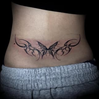 Symmetrical ornamental butterfly tattoo with flowing vine-like wings spanning across the lower back in black ink.