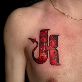 Red and black lettering tattoo with floral and figure details on the upper chest of a shirtless person wearing a gold chain.