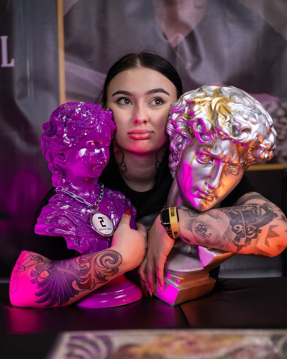 Woman displaying elaborate wing and ornamental tattoos on both arms in purple and orange lighting, posing with decorative sculptural heads.