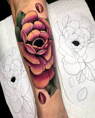 Colorful peony tattoo on forearm featuring purple, pink, and gold petals with green leaves, accompanied by line-drawn design references on white paper.