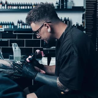 Tattoo artist wearing glasses and black gloves applies ink to a client's arm in a studio with shelves of ink bottles visible in the background.