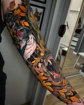 Colorful bird and animal sleeve tattoo featuring orange autumn leaves, a woodpecker, owl, and bear with detailed facial features on the forearm and upper arm.