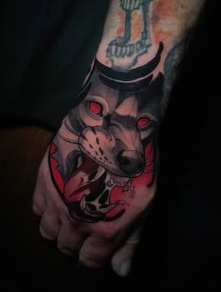 Demon face tattoo with red eyes and black markings on the back of a hand, featuring horns and sharp teeth details.