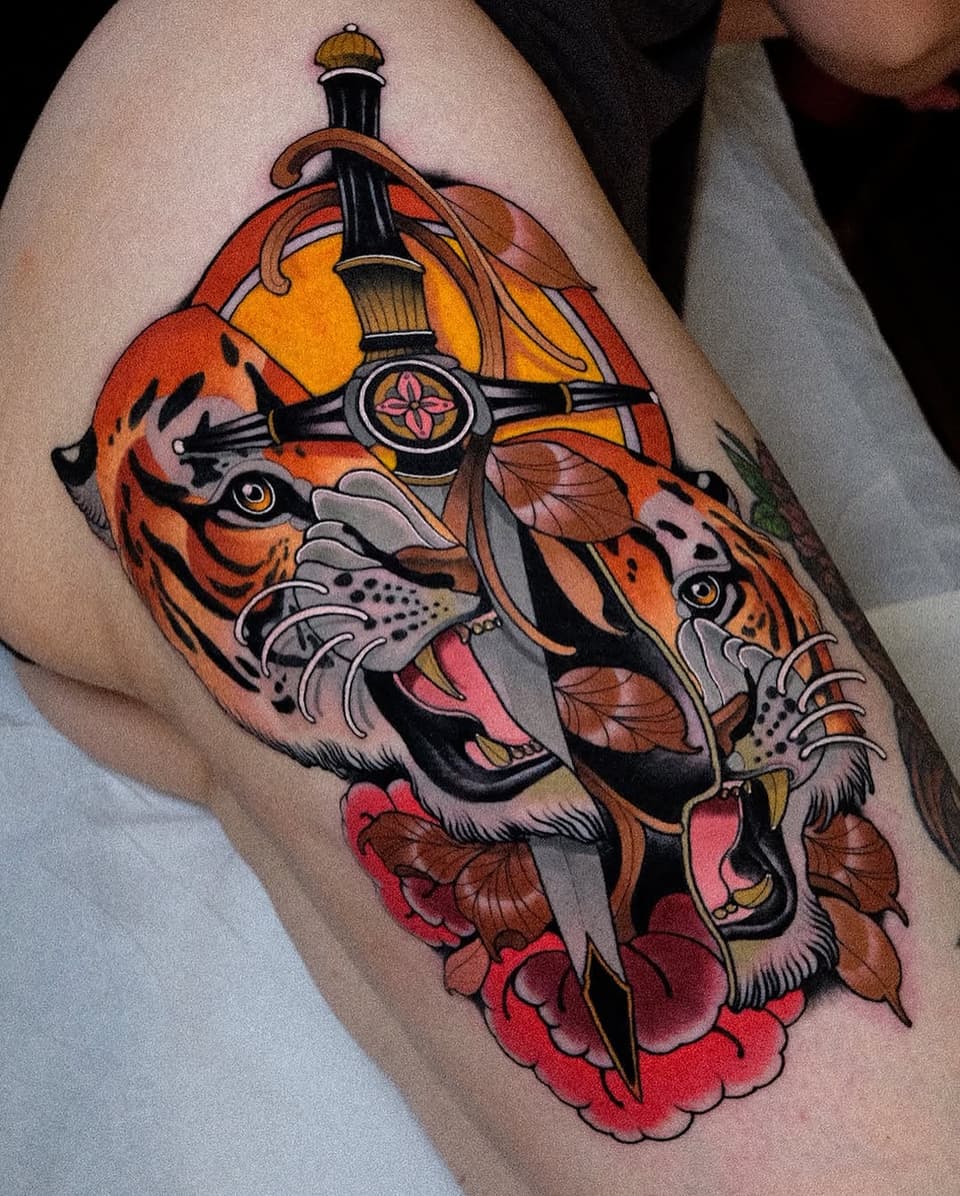 Tiger head with sword piercing through it, surrounded by red flowers and autumn leaves, tattooed on upper arm in orange, red, black, and yellow colors.