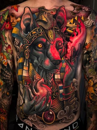 Large-scale back tattoo featuring an Egyptian jackal deity head with golden ornaments, surrounded by colorful flowers, pyramids, and decorative elements in red, gold, and black tones.