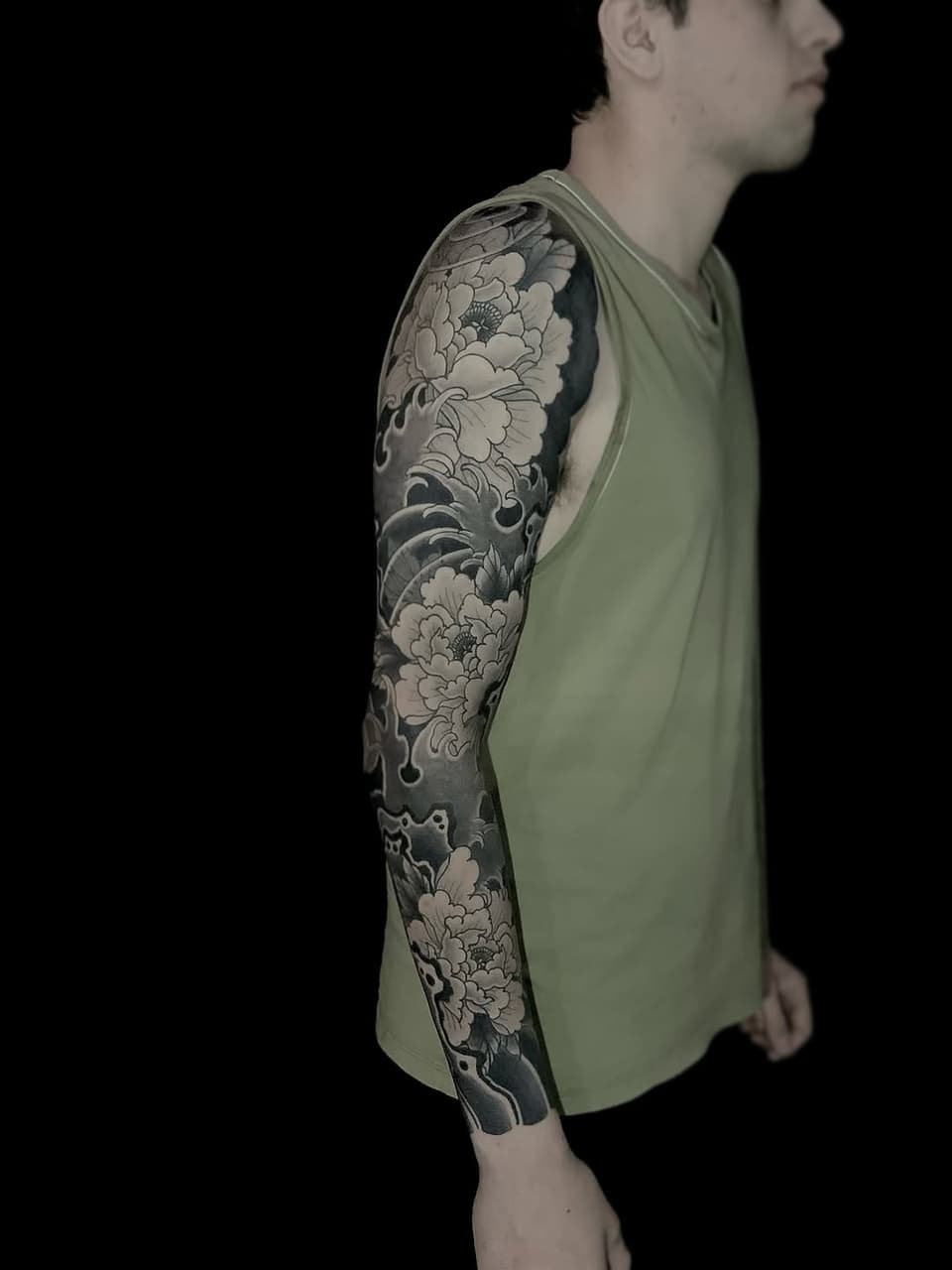 Man wearing a light green sleeveless shirt displaying a full arm sleeve tattoo featuring large detailed flowers and ornamental scrollwork in black and gray ink.
