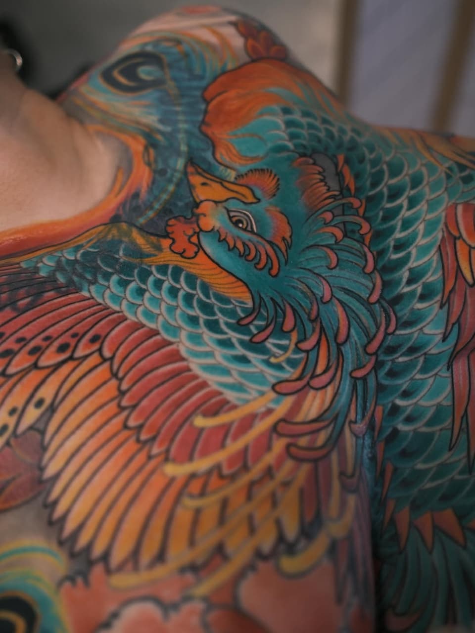 Colorful phoenix tattoo on shoulder and chest featuring orange, teal, and red plumage with detailed feather work and ornamental details.