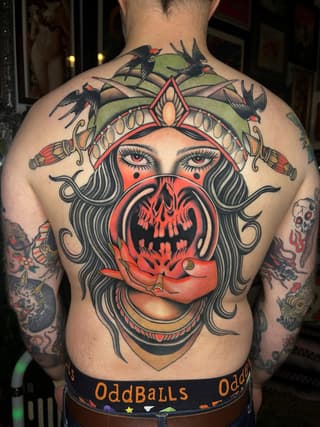 Large-scale back tattoo featuring a female face with red ornamental headdress, skeletal mouth, swallows, and decorative elements in red, black, and green.