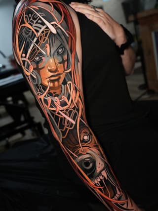 Full-sleeve arm tattoo featuring a woman's face with ornate decorative elements in orange, red, and black, with a skull design at the wrist.