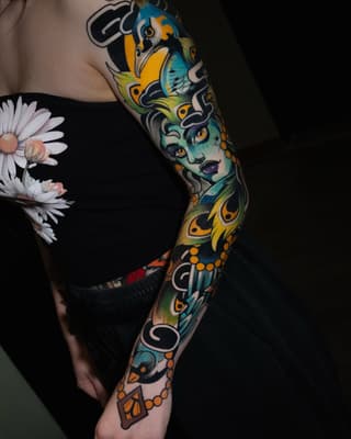 Full-sleeve arm tattoo featuring a turquoise-faced woman with yellow and black ornamental patterns, birds, and decorative elements in blue, yellow, orange, and black.