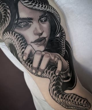 Detailed monochromatic shoulder tattoo featuring a woman's face with serpent motifs wrapping around her neck and shoulder, adorned with jewelry details.