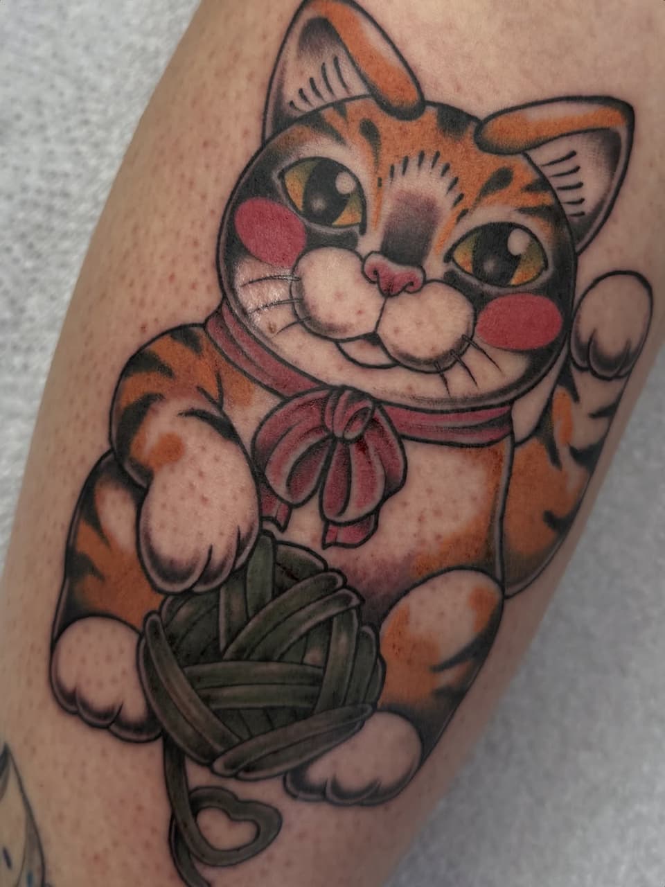 Maneki-neko lucky cat tattoo on forearm featuring orange tabby with red cheeks, white muzzle, red collar bow, and green basket, in orange, red, black, and cream colors.