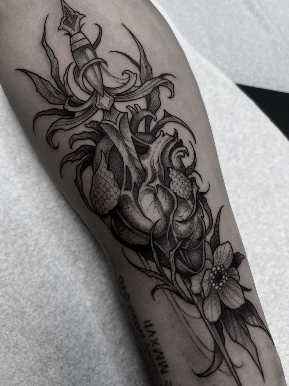 Anatomical heart with dagger, ornate scrollwork, and flowering plants in black ink on forearm, with small text visible near wrist.