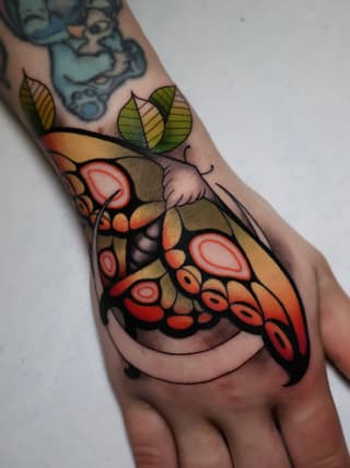 Colorful butterfly tattoo on the hand featuring red and orange wings with green leaves, black outlines, and detailed wing patterns.