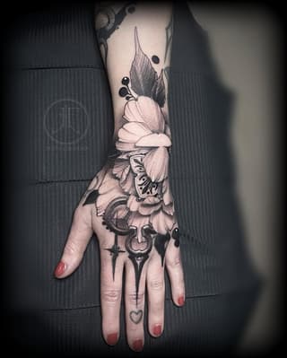 Hand and forearm tattoo featuring flowers, butterflies, moons, and decorative symbols in black ink on pale skin, displayed against dark fabric.