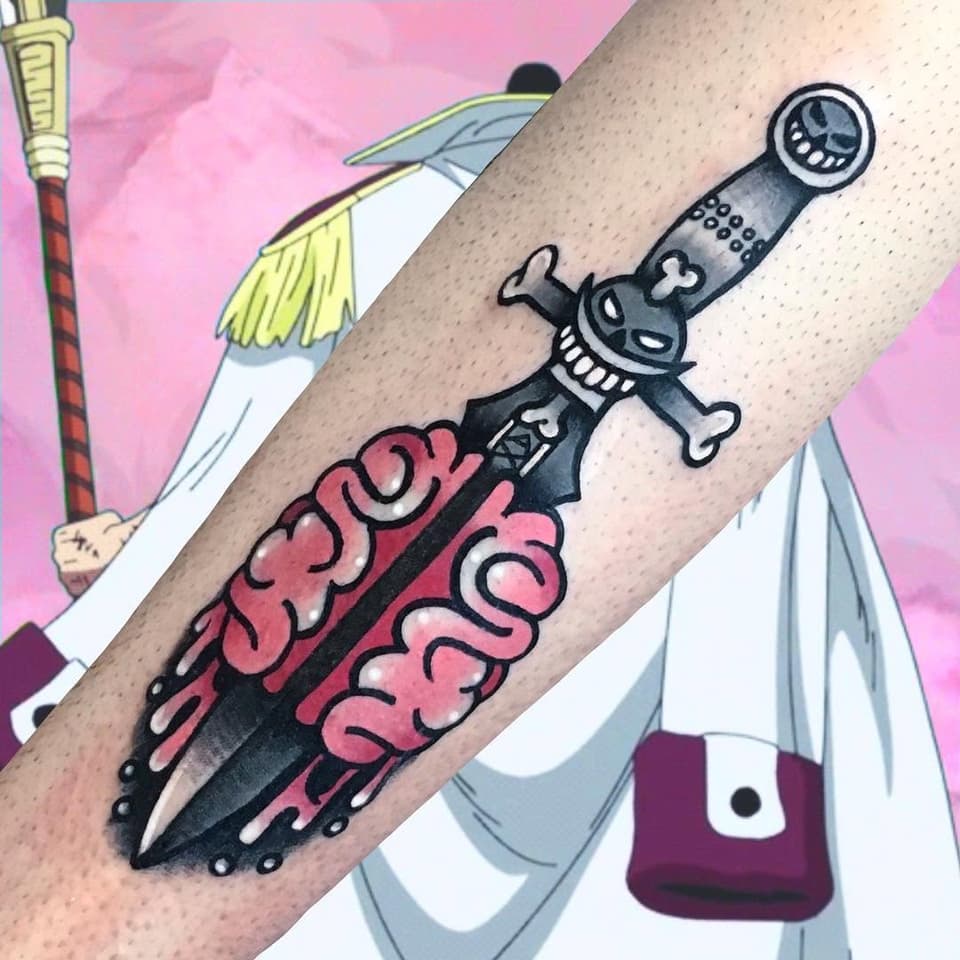 Tattoo on forearm featuring a black skeletal sword with pink ornamental scrollwork, topped with a grinning skull character, displayed against colorful background illustrations.