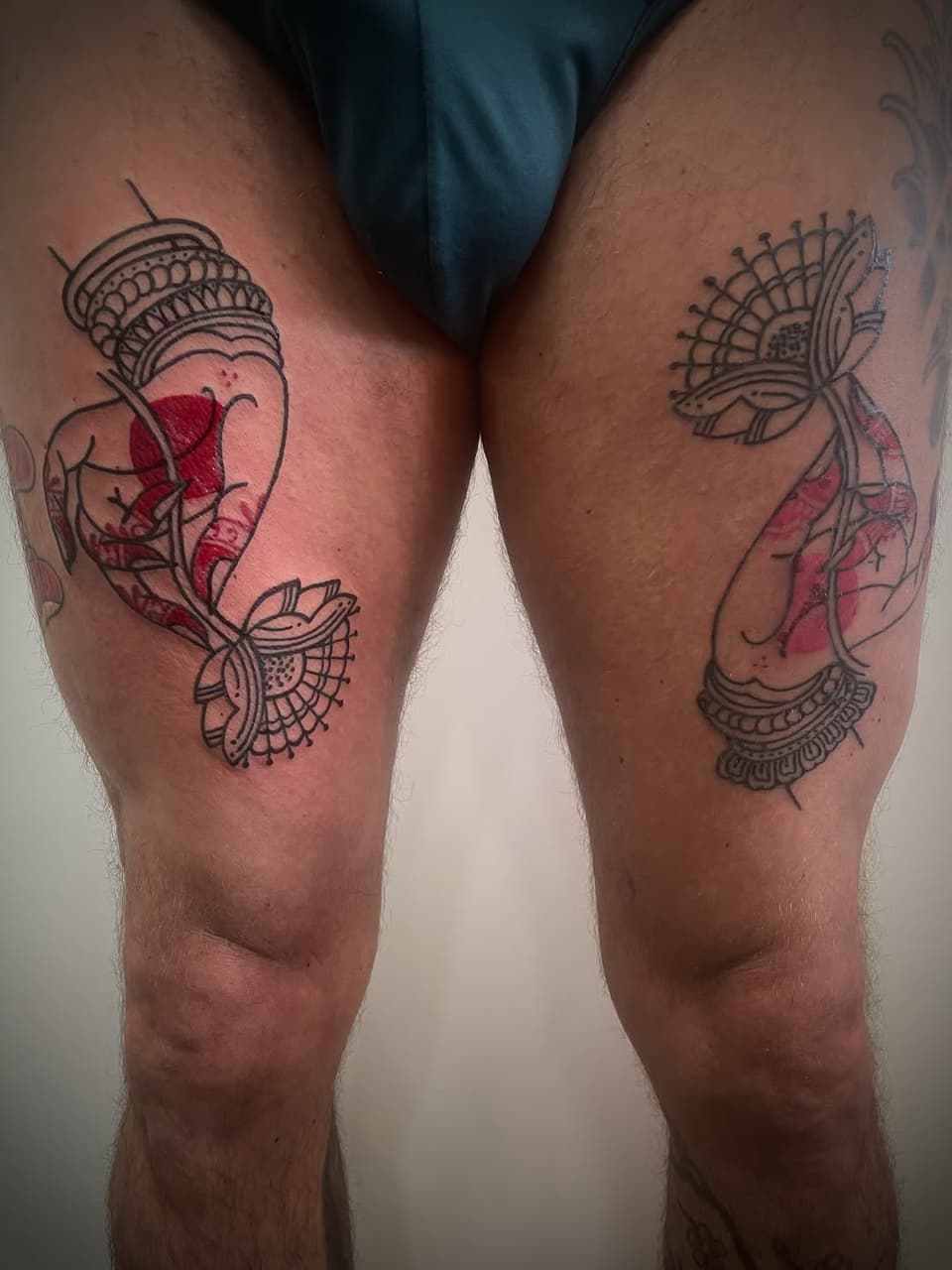 Matching leg tattoos featuring ornate fan designs with red watercolor accents and decorative linework on both thighs.
