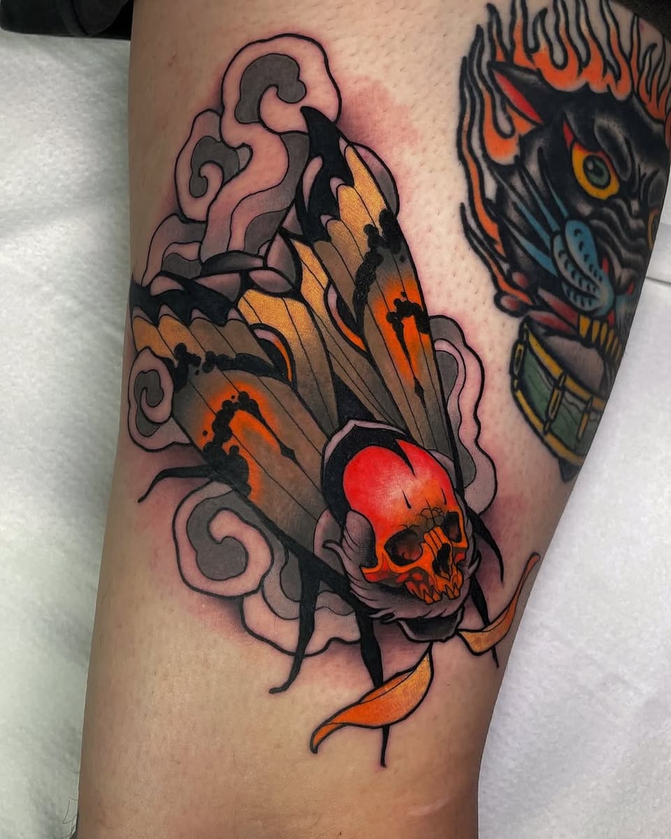 Colorful arm tattoo featuring bat wings with a red skull, orange and tan shading, pink swirling clouds, and black outlines on skin.