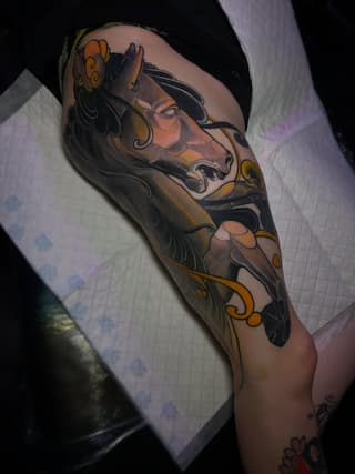 Decorative horse head tattoo on upper arm featuring black linework with gold and orange accents, ornamental flourishes, and flowing mane details.