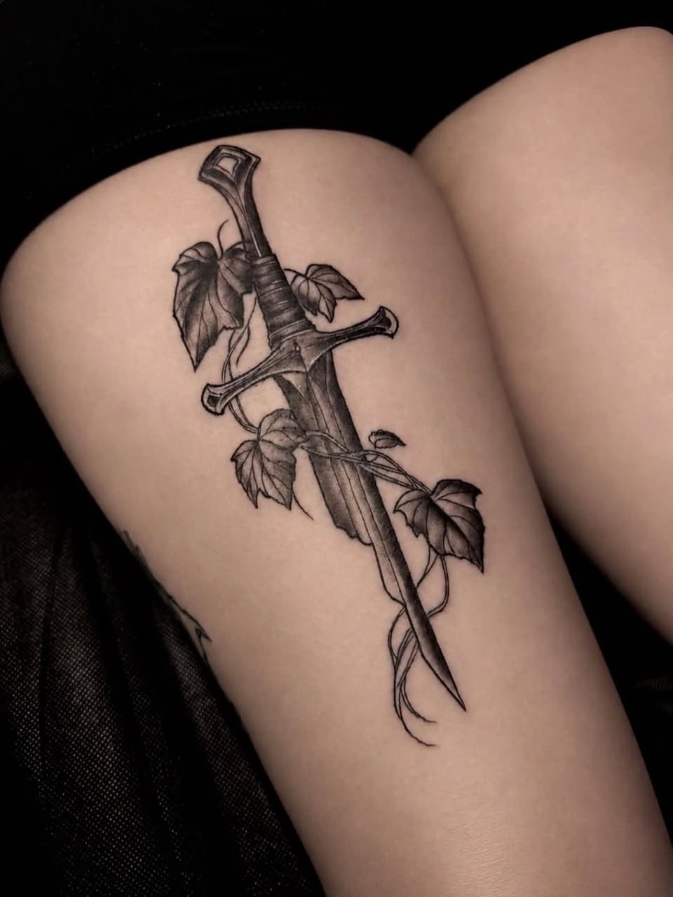 Sword with ornate cross-guard and wrapped handle adorned with hop plant leaves and vines, tattooed on thigh in black ink.
