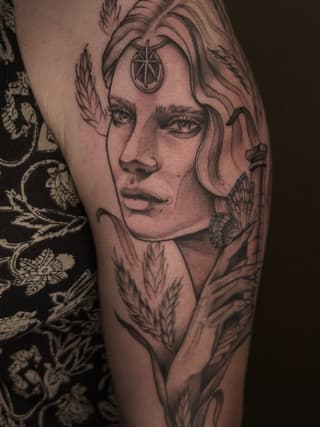 Portrait tattoo on upper arm featuring a face with closed eyes, ornamental third eye symbol on forehead, surrounded by feathered and botanical elements in black ink.