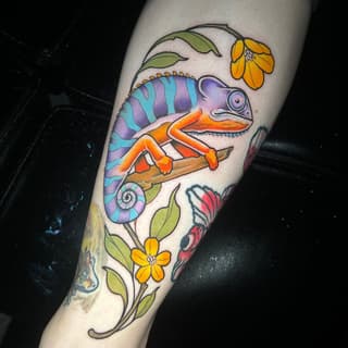 Colorful chameleon perched on a branch with purple, turquoise, and orange coloring, surrounded by yellow flowers and green leaves, tattooed on the calf.