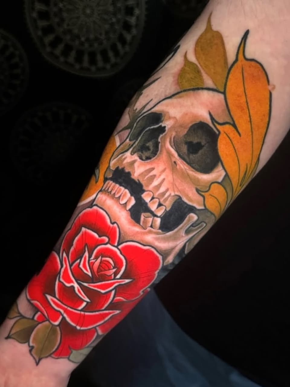 Skull with orange leaves and red rose tattoo on forearm, featuring detailed shading in cream, black, and red colors.