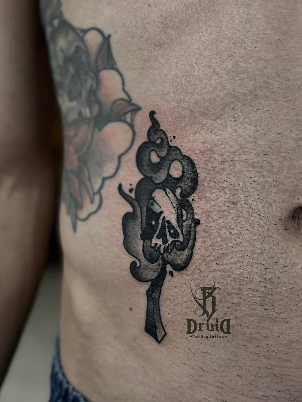 Black ink tattoo on the ribs featuring intertwined snakes with a skull, accompanied by a dagger and artist's logo and text below.