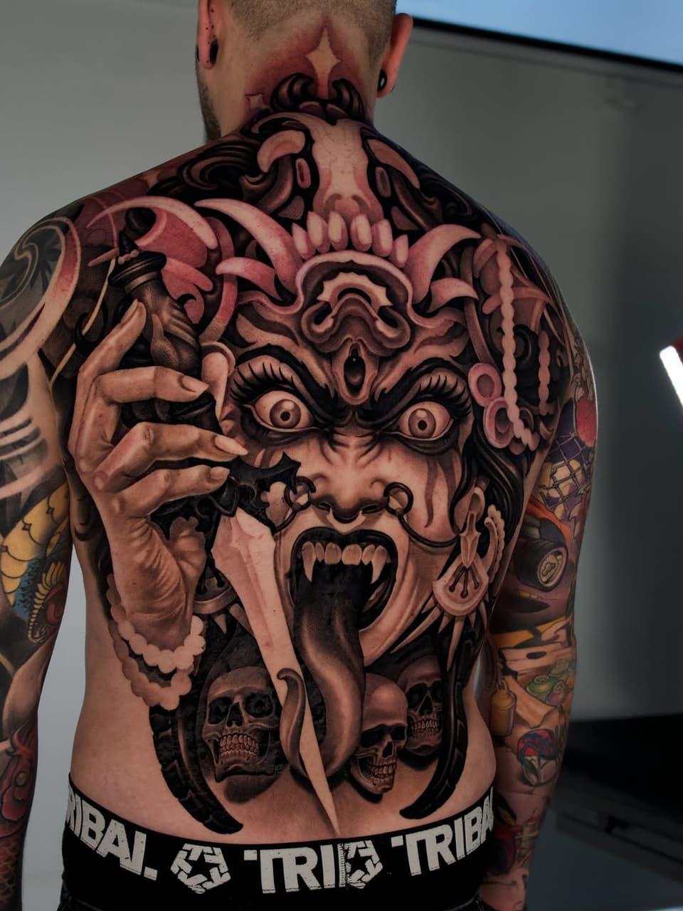 Back tattoo featuring a demonic face with elaborate horns, wide eyes, and open mouth, with skulls and ornamental details below, rendered in red and black ink.